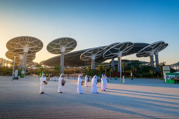 People in traditional attire walk in front of futuristic, large solar panel structures under a clear blue sky.