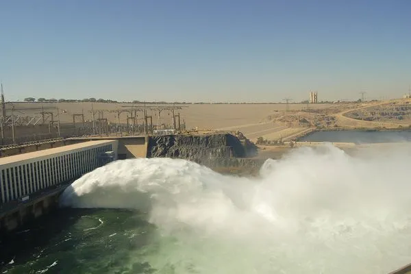 Water gushing from the Aswan High Dam into the Nile River, with arid desert landscape and clear blue sky in the background.