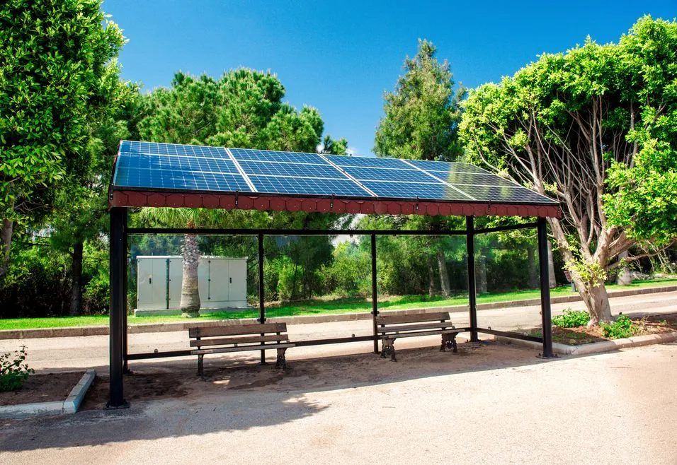 Bus stop with solar panels on the roof, surrounded by trees and greenery under a clear blue sky.