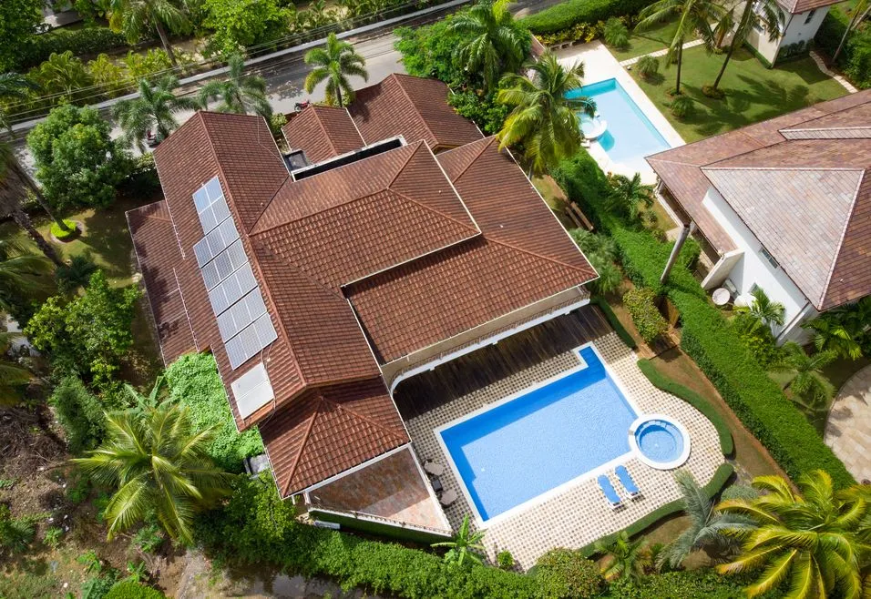 Aerial view of a large house with a red-tiled roof, solar panels, a swimming pool, and lush greenery surrounding the property.