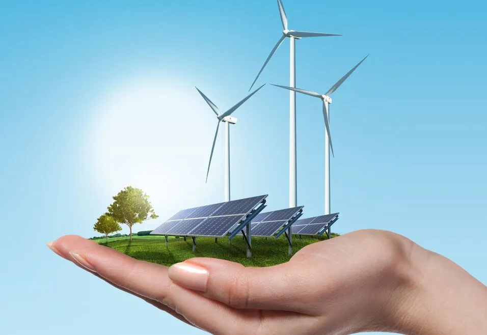 A hand holding a miniature landscape with solar panels, wind turbines, and a tree, symbolizing renewable energy under a clear blue sky.